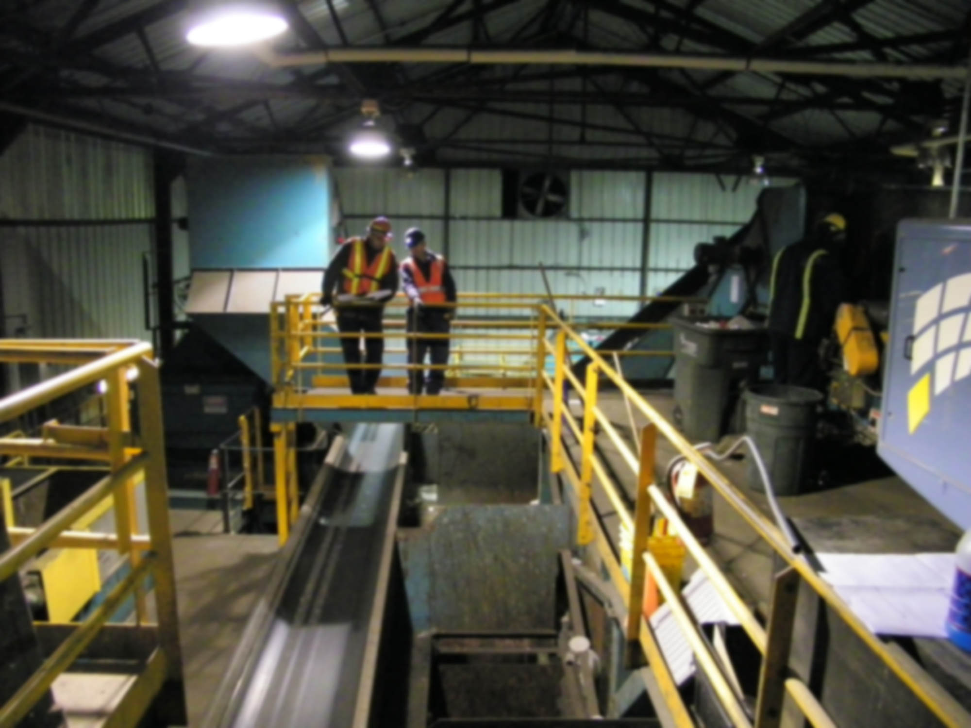 Two crew members in safety vests reviewing plans on a catwalk above conveyors in a recycling plant