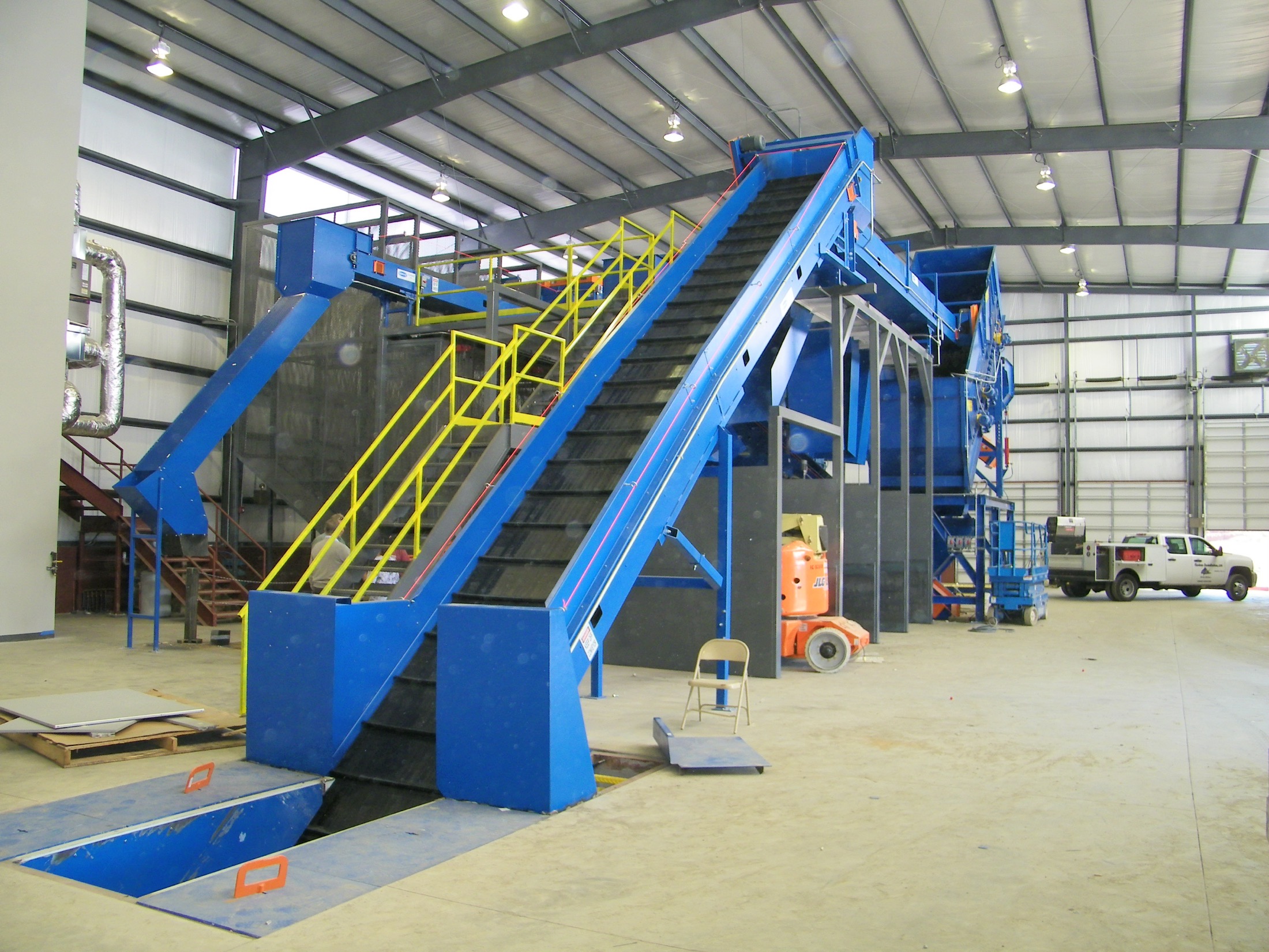 A large blue Remcon conveyor and sorting system with an incline feed conveyor and access stairs, installed in a recycling facility